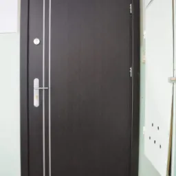 Ciemnobrązowe skrzydło z dwoma pionowymi, srebrnymi paskami, zamontowane w ościeżnicy, widoczny zamek i klamka na tle jasnej ściany korytarza.