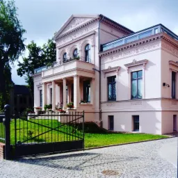 Elegancka, dwupiętrowa willa w pastelowym różu z kolumnami, balkonem i ozdobnymi detalami architektonicznymi, otoczona zadbanym trawnikiem i kostką brukową, widziana zza czarnej, metalowej bramy.