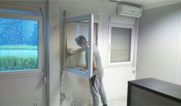 Mężczyzna w szarym dresie czyści wewnętrzną stronę otwartego okna w biurze, z widokiem na zewnątrz na zielone pole i deszczowy dzień; w tle widoczne okno z żaluzjami i klimatyzator.