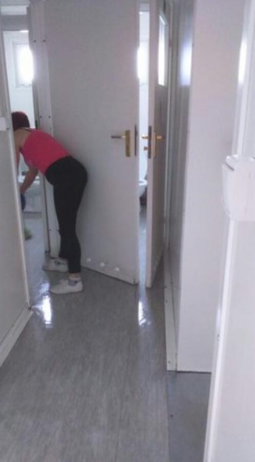 Kobieta w różowej koszulce i czarnych legginsach czyści podłogę w wąskim, białym korytarzu przy otwartych drzwiach do łazienki, widoczna toaleta.