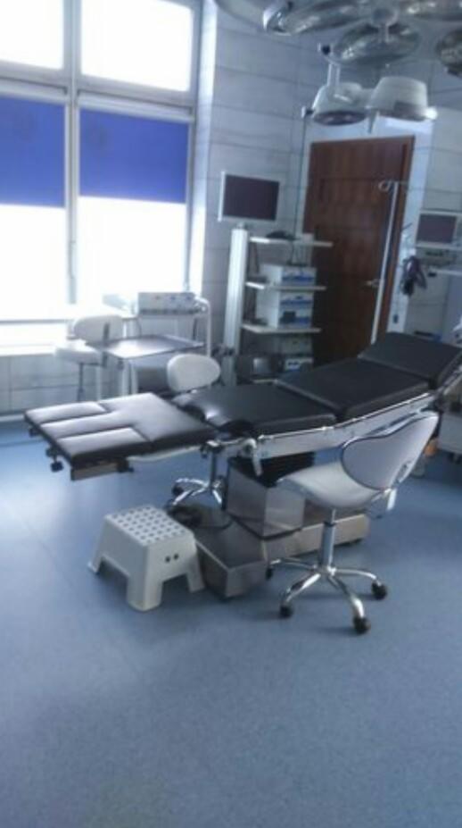 Sala operacyjna z nowoczesnym stołem operacyjnym, krzesłem na kółkach i białym stołkiem, oświetlona przez lampy sufitowe, z oknami z niebieskimi roletami w tle.