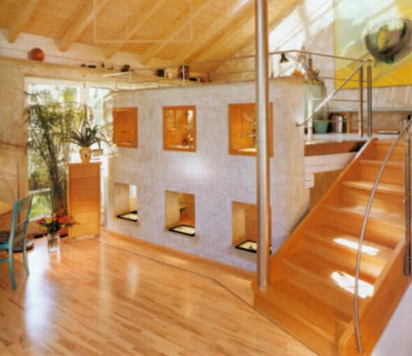 Jasne, przestronne wnętrze z drewnianą podłogą, schodami z metalową poręczą i ścianą z wnękami oraz oknami, roślina doniczkowa przy oknie, widoczne elementy wystroju wnętrza w stylu skandynawskim.