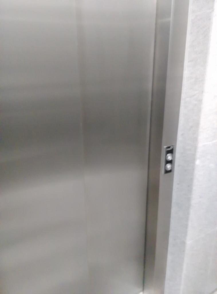 Metalowe drzwi windy z przyciskami w pionowym panelu, widoczne fragmenty marmurowej ściany.