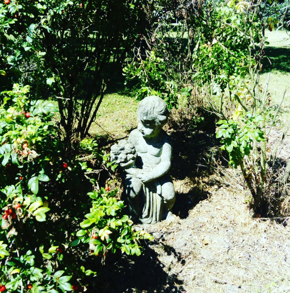 Kamienna figurka dziecka z kwiatami w przysiadzie, otoczona krzewami z czerwonymi owocami i suchą ściółką w ogrodzie.