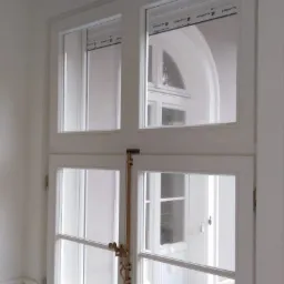 Białe, dwuskrzydłowe okno z mosiężnym zamknięciem na tle klasycznego wnętrza z dekoracyjnym sufitem i widokiem na białe drzwi balkonowe.