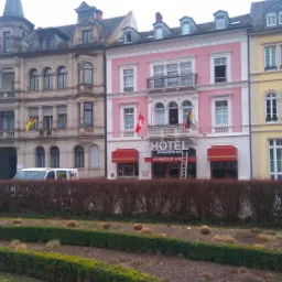 Fasady kolorowych kamienic z oknami, w tym różowego hotelu ze schodami opartymi o wejście, widoczne zza niskiego żywopłotu.
