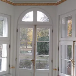 Wnętrze jasnego pokoju z białymi, przeszklonymi drzwiami balkonowymi z półokrągłym nadświetlem i złotymi okuciami, widok na park przez okna, dekoracyjny złoty ornament na suficie, podłoga w trakcie...