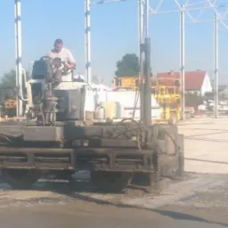 Wylewka posadzki przemysłowej za pomocą specjalistycznej maszyny. Operator kontroluje proces wylewania betonu na dużej powierzchni pod zadaszeniem konstrukcji stalowej.