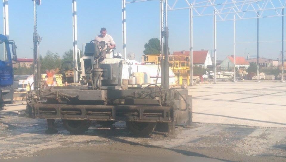 Wylewka posadzki przemysłowej za pomocą specjalistycznej maszyny. Operator kontroluje proces wylewania betonu na dużej powierzchni pod zadaszeniem konstrukcji stalowej.