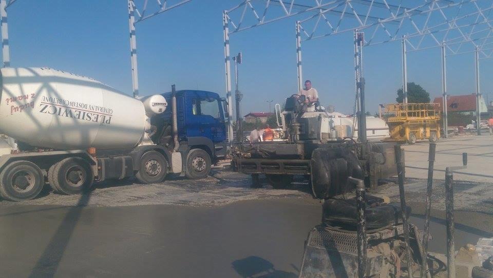 Betoniarka i maszyna do wylewania betonu na budowie hali. Widoczny operator maszyny i konstrukcja stalowa.