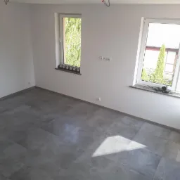 Puste pomieszczenie z szarymi płytkami podłogowymi, białymi ścianami i dwoma oknami z widokiem na zieleń na zewnątrz. Na jednym z parapetów leży czarny sprzęt elektroniczny.