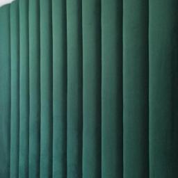 Usługi remontowo-budowlane Łukasz Cywiński - Zielony, tapicerowany panel ścienny z pionowymi przeszyciami, tworzący wezgłowie łóżka, widoczny fragment białej pościeli w dolnym rogu.