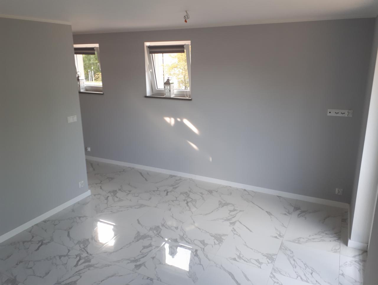 Pusty pokój z szarymi ścianami i marmurową podłogą, dwa okna z lampionami na parapetach, widoczne gniazdka elektryczne na ścianie.