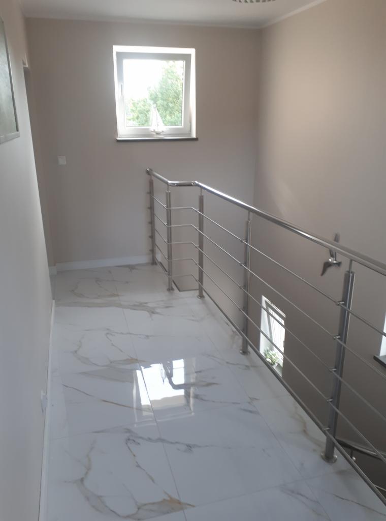 Wnętrze klatki schodowej z marmurową posadzką i nowoczesną, metalową balustradą. Okno z widokiem na zieleń w tle.