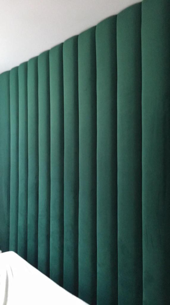 Zielony, tapicerowany panel ścienny z pionowymi przeszyciami, tworzący wezgłowie łóżka, widoczny fragment białej pościeli w dolnym rogu.