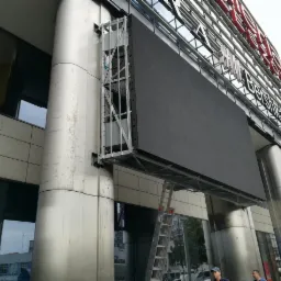 Duży ekran LED zamontowany na metalowej konstrukcji przy budynku centrum handlowego, widoczna drabina i pracownicy na dole.