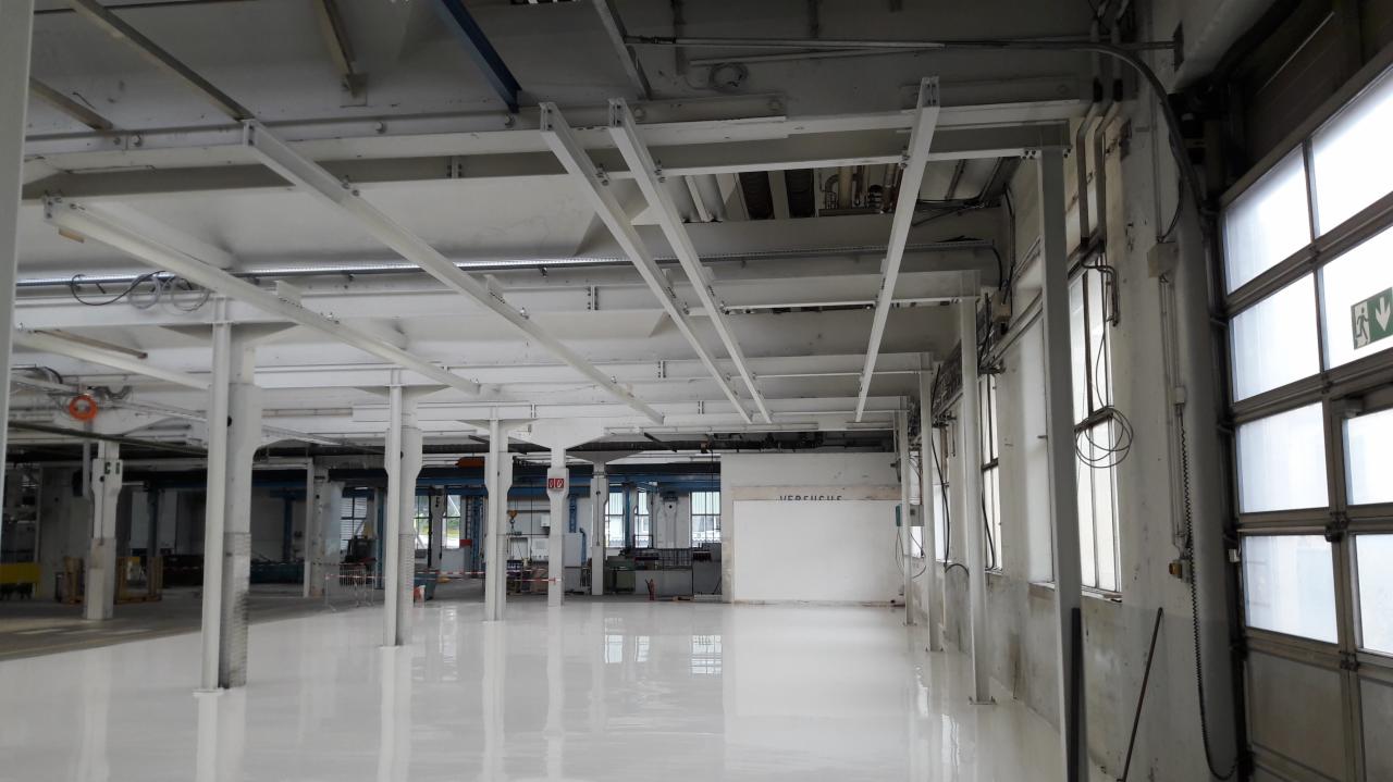 Wnętrze hali produkcyjnej z białymi stalowymi słupami i belkami podtrzymującymi sufit, świeżo wylana biała posadzka epoksydowa, duże okna w tle, widoczne instalacje pod sufitem.