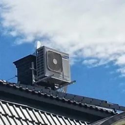 Klimatyzator zewnętrzny na dachu domu pod błękitnym niebem z chmurami. Montaż na specjalnej konstrukcji wsporczej. Widok z dołu, dach&oacute;wka w pierwszym planie.