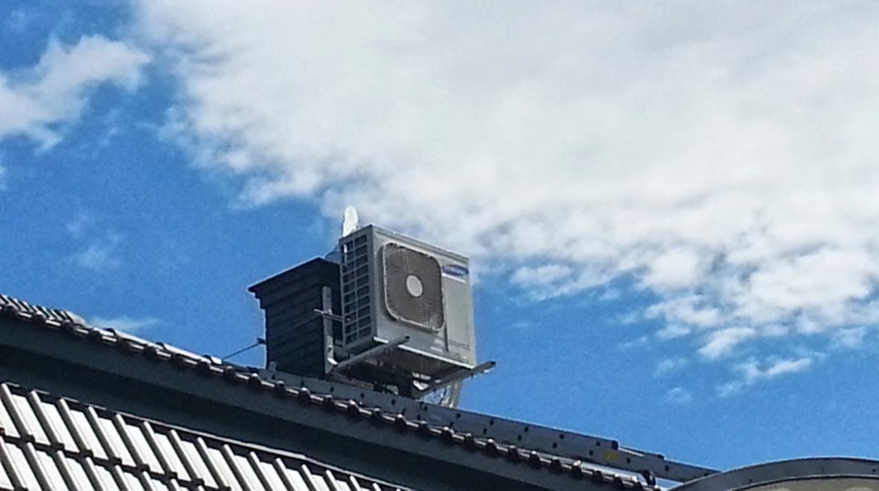 Klimatyzator Samsung zamontowany na dachu budynku, widoczny z dołu na tle błękitnego nieba z chmurami. Instalacja na wspornikach, obok komin wentylacyjny.