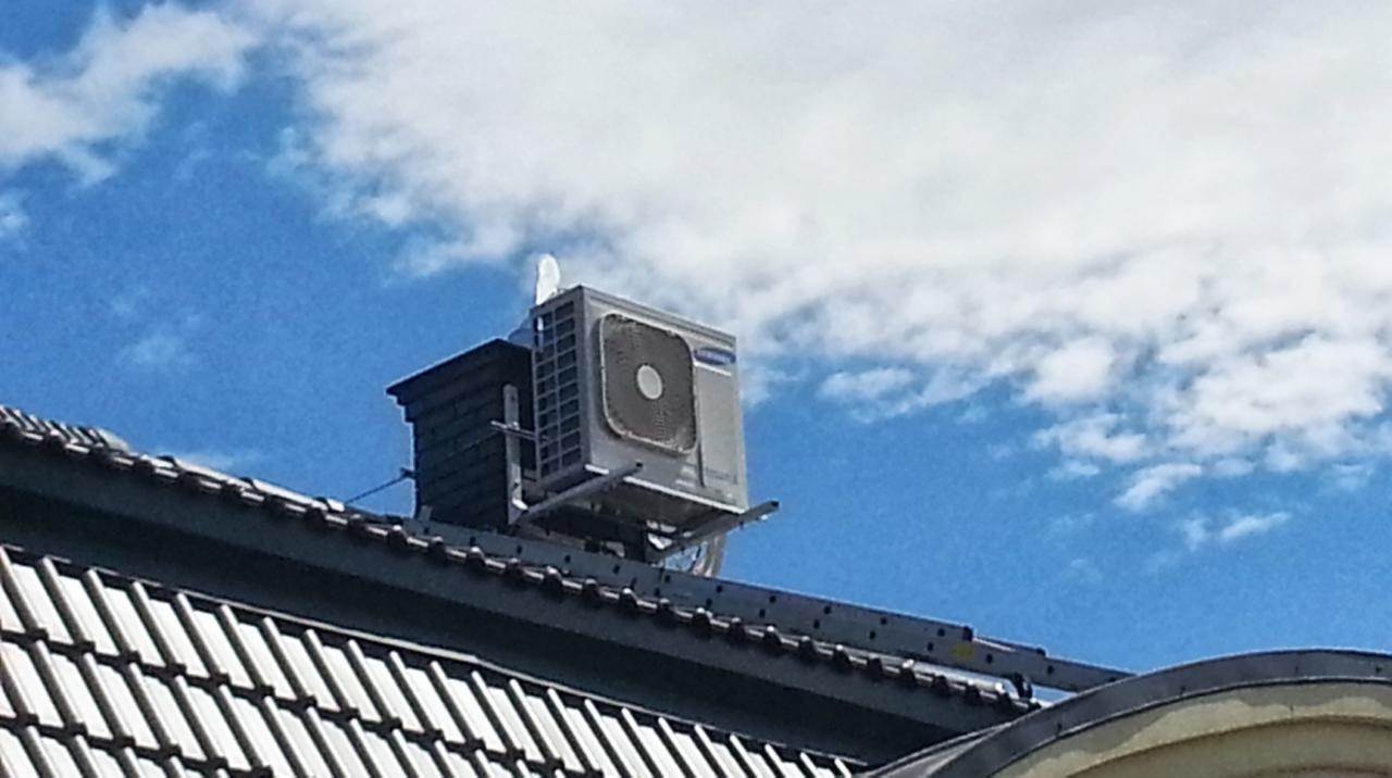 Klimatyzator zewnętrzny na dachu domu pod błękitnym niebem z chmurami. Montaż na specjalnej konstrukcji wsporczej. Widok z dołu, dachówka w pierwszym planie.