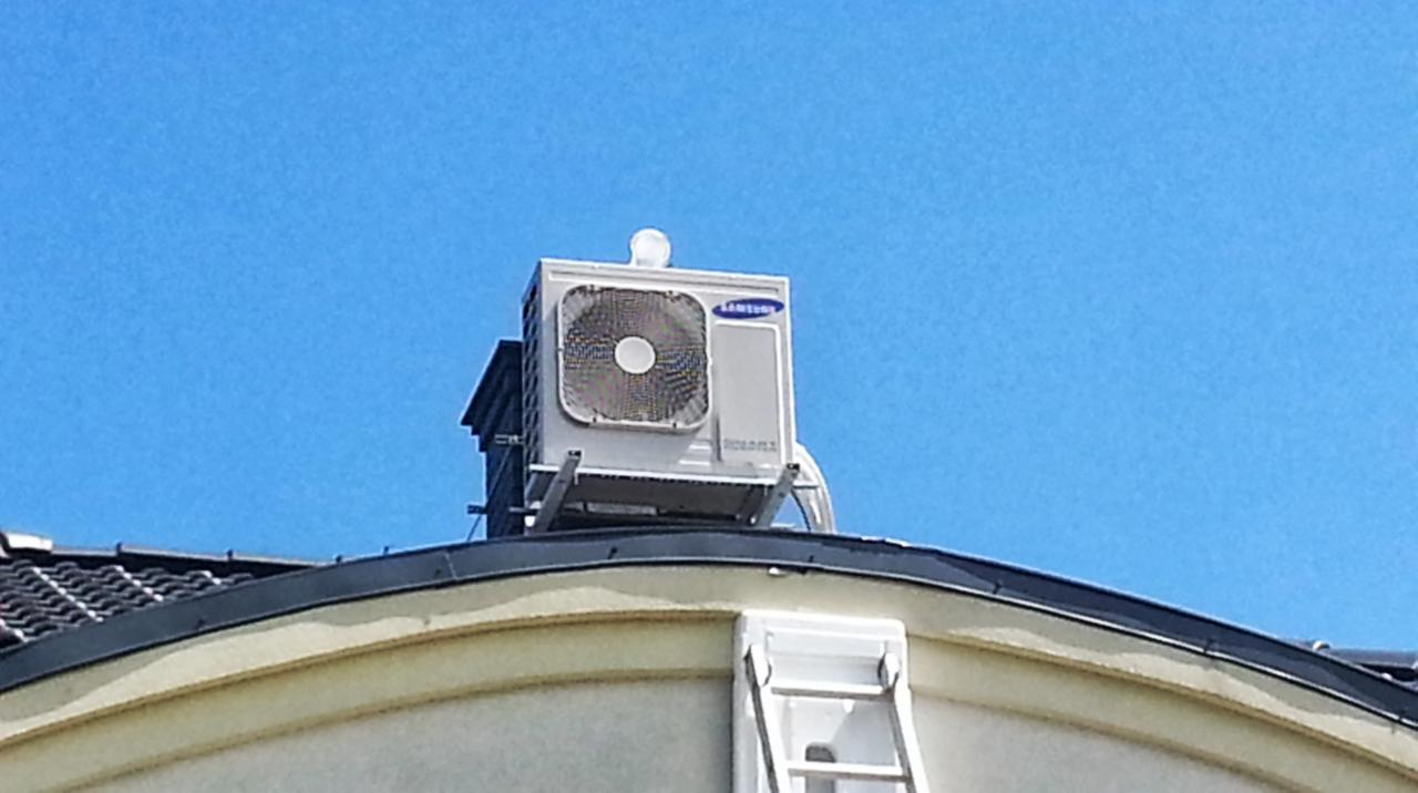 Biała jednostka klimatyzacji Samsung zamontowana na dachu domu, widoczna drabina i błękitne niebo w tle. Instalacja na metalowej konstrukcji.