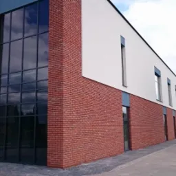 Budynek z ceglaną elewacją i dużymi oknami. Widoczne trzy małe okna z zamontowanymi roletami zewnętrznymi. Ujęcie z zewnątrz, w tle zieleń i błękitne niebo.