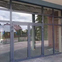 Szklana fasada budynku z szarą ramą, widok z zewnątrz. W szybach odbija się krajobraz, a po bokach widać fragmenty ceglanej ściany.