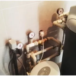 Zamontowany zmiękczacz wody z manometrami i rurami w łazience wyłożonej jasnymi płytkami, widoczny wystający kabel zasilający.