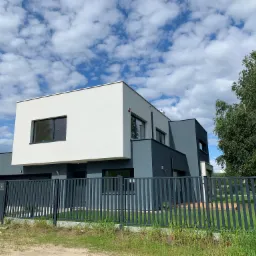 Dwukondygnacyjny dom w nowoczesnym stylu, z białym i szarym wykończeniem elewacji, czarnymi oknami, ogrodzony metalowym płotem na tle błękitnego nieba z chmurami.