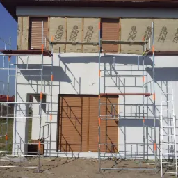 Dom w trakcie ocieplania, widoczne rusztowania i płyty izolacyjne Strotex 1300. Częściowo zasłonięte okna roletami zewnętrznymi. Słoneczny dzień.