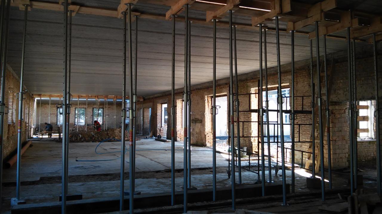 Wnętrze budynku w trakcie budowy, z widocznymi podporami stropowymi, ceglanymi ścianami i oknami. W tle pracują dwie osoby.