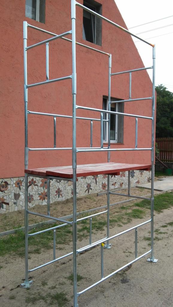 Metalowe rusztowanie na tle pomarańczowej ściany budynku z oknami. Widoczna mozaika kwiatowa na fundamencie. Ujęcie z dołu, lekko z prawej strony.