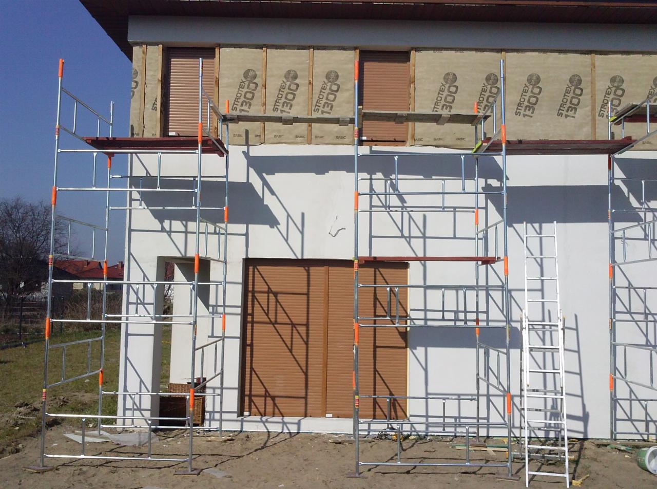 Dom w trakcie ocieplania, widoczne rusztowania i płyty izolacyjne Strotex 1300. Częściowo zasłonięte okna roletami zewnętrznymi. Słoneczny dzień.