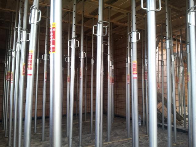 Szereg stalowych podpór budowlanych, rozmieszczonych gęsto na betonowej posadzce, wspierających drewniany strop. Widoczne czerwone etykiety na podporach.