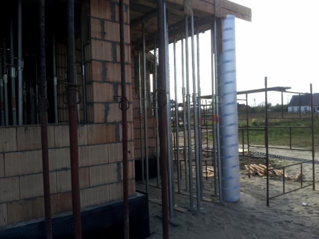 Wznoszenie ściany z pustaków ceramicznych, widoczne podpory budowlane i rusztowanie. Słup zabezpieczony folią. Budowa w toku, widok na okoliczne zabudowania.