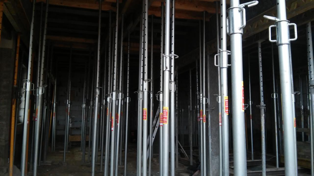 Wnętrze konstrukcji szalunkowej z licznymi, metalowymi stemplami budowlanymi podtrzymującymi strop. Widoczne elementy drewniane szalunku i drabina w tle.