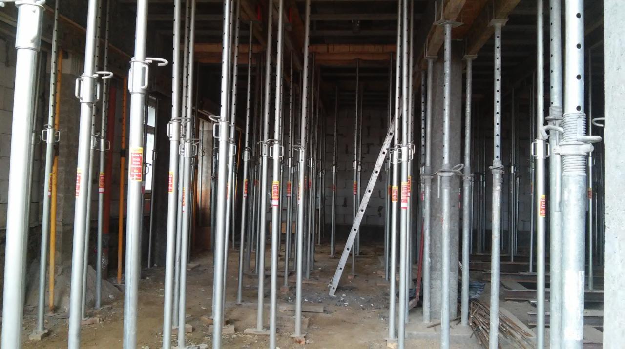 Wnętrze w trakcie budowy: liczne stalowe stemple podtrzymują strop, widoczna drabina i ściany z pustaków. Etap prac przed wylaniem betonu.