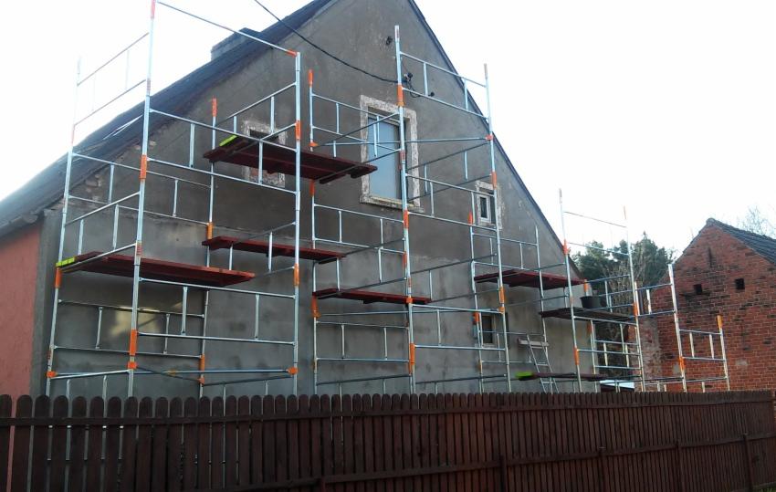 Szarej elewacji domu w trakcie renowacji towarzyszą metalowe rusztowania z drewnianymi podestami, widoczne zza drewnianego płotu.