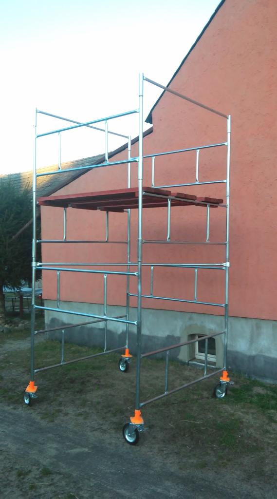 Stalowe rusztowanie na kółkach z drewnianym podestem, ustawione przed budynkiem z pomarańczową elewacją i małym okienkiem w fundamencie.