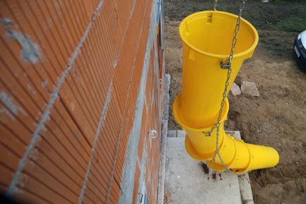 Ściana z czerwonej cegły dziurawki z zamontowanym żółtym zsypem budowlanym, wiszącym na łańcuchach. Widok z boku, częściowo widoczny samochód w tle.