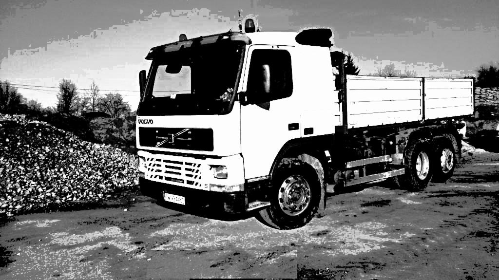 Biały samochód ciężarowy Volvo z otwartą skrzynią ładunkową na błotnistym terenie, obok sterty żwiru, w czarno-białej tonacji.