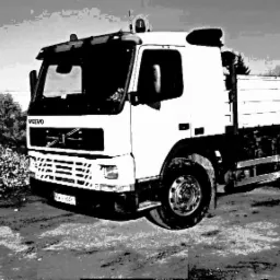 Czarno-białe zdjęcie białego samochodu ciężarowego Volvo z otwartą skrzynią ładunkową na tle hałdy gruzu i drzew. Widoczne błotniste drogi i fragment tablicy rejestracyjnej.