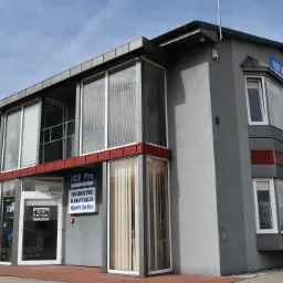 Budynek firmy ICE Pro z szarymi ścianami, dużymi oknami i szyldem informującym o ofercie: chłodnictwo, klimatyzacja, pompy ciepła. Widoczne godziny otwarcia na drzwiach wejściowych.