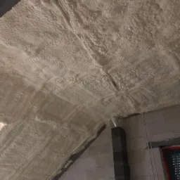 Poddasze po izolacji natryskowej, widoczne nierównomierne pokrycie pianką na skośnych i poziomych powierzchniach, z elementami instalacji elektrycznej wystającymi spod izolacji.