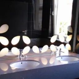 Nowoczesna łazienka z dwoma umywalkami wpuszczonymi w błyszczący, ciemny blat, oświetlona lampami w kształcie kropli i odbiciem w lustrze na tle czarnych płytek.