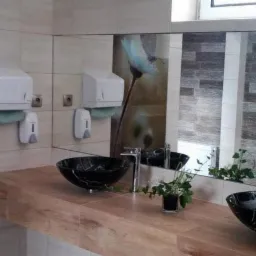 Nowoczesna łazienka publiczna z drewnianym blatem, czarnymi umywalkami w kształcie misy, dwoma dozownikami na papierowe ręczniki i dużym lustrem z dekoracyjnym panelem imitującym kamień i motywem...