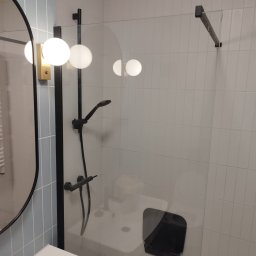 Gładko.pl - Nowoczesna łazienka z czarną armaturą, szklaną kabiną prysznicową i okrągłym lustrem z oświetleniem kulowym, wykończona płytkami w odcieniach bieli i błękitu.