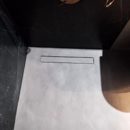 Gładko.pl - Nowoczesna kabina prysznicowa z szarym wykończeniem podłogi, czarnymi ścianami imitującymi marmur, odpływem liniowym i miedzianą armaturą, widoczne stopy w skarpetkach.