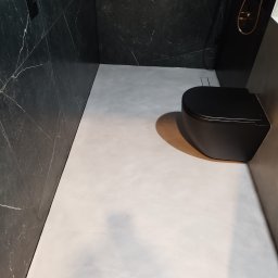 Gładko.pl - Nowoczesna łazienka z czarną armaturą i szarym mikrocementem na podłodze, ściany wykończone czarnym marmurem z białymi żyłami.