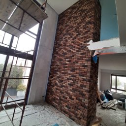 Gładko.pl - Wnętrze w trakcie prac: częściowo wykończona ściana z cegieł dekoracyjnych, rusztowanie przy oknie, zabezpieczona folią podłoga i widoczne materiały budowlane.